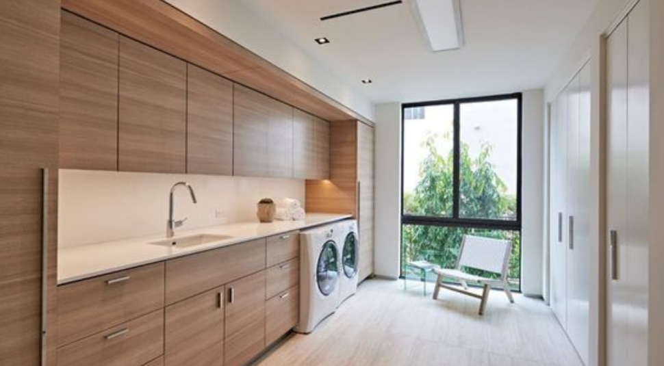 A laundry room fitted with two front loading wachine machines and with wooden finish vanity cabinets. The room comes with a big glass window and has a reading table and chair.