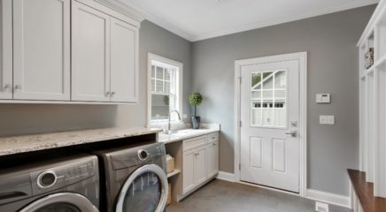 A grey color wall themed laundry room fitted with white cupboards, white door, white window. It is also fitted with two grey coloured front loading washing machines.