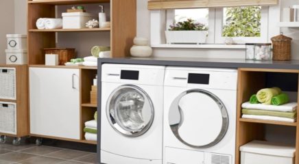 A white themed laundry room fitted with two white front load washing machine and dryer. Also, fitted with wooden cupboard and storage baskets.