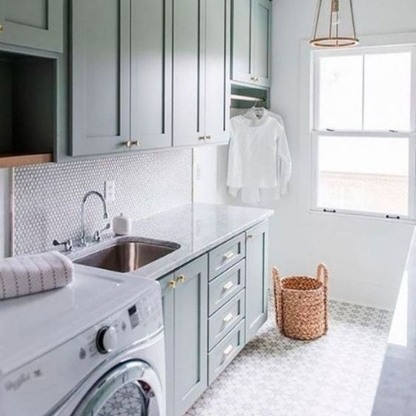 A white and off blue themed laundry room fitted with a white front load washing machine. It is well lit & ventilated, big windows and lights on the ceiling. There is a laundry basket and storage cupboards